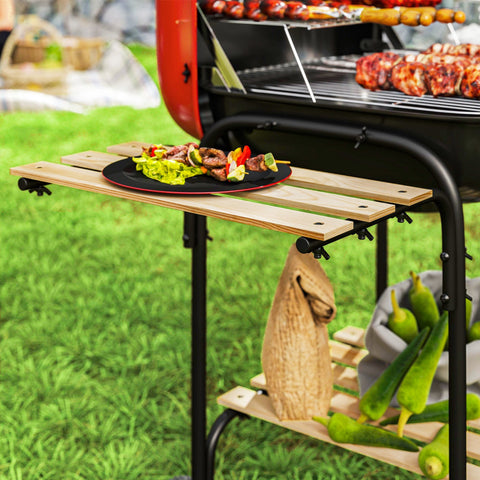 A rustic wooden outdoor grill cart with black metal legs holding fresh okra beside a red portable bbq with grill grate and warming rack