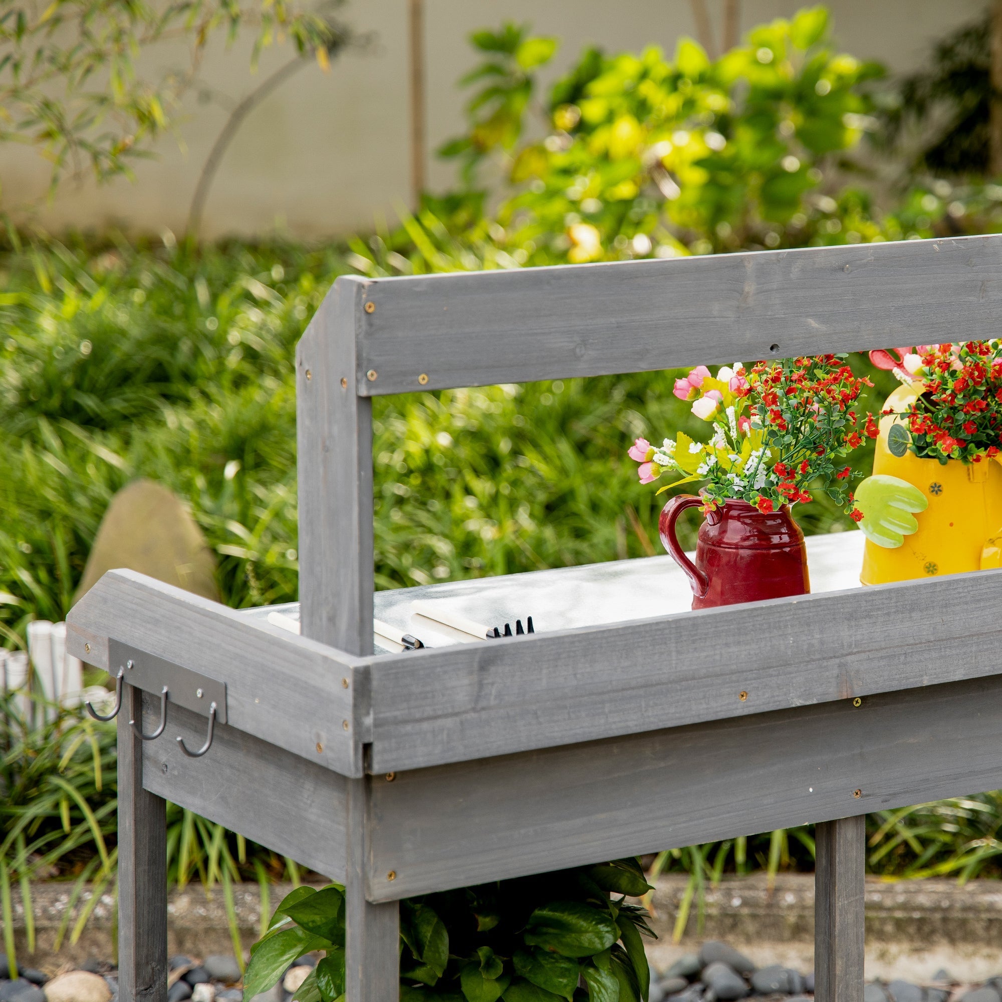 Gray wooden garden potting table with galvanized metal top, hooks, drawer, and red watering can beside flower pot