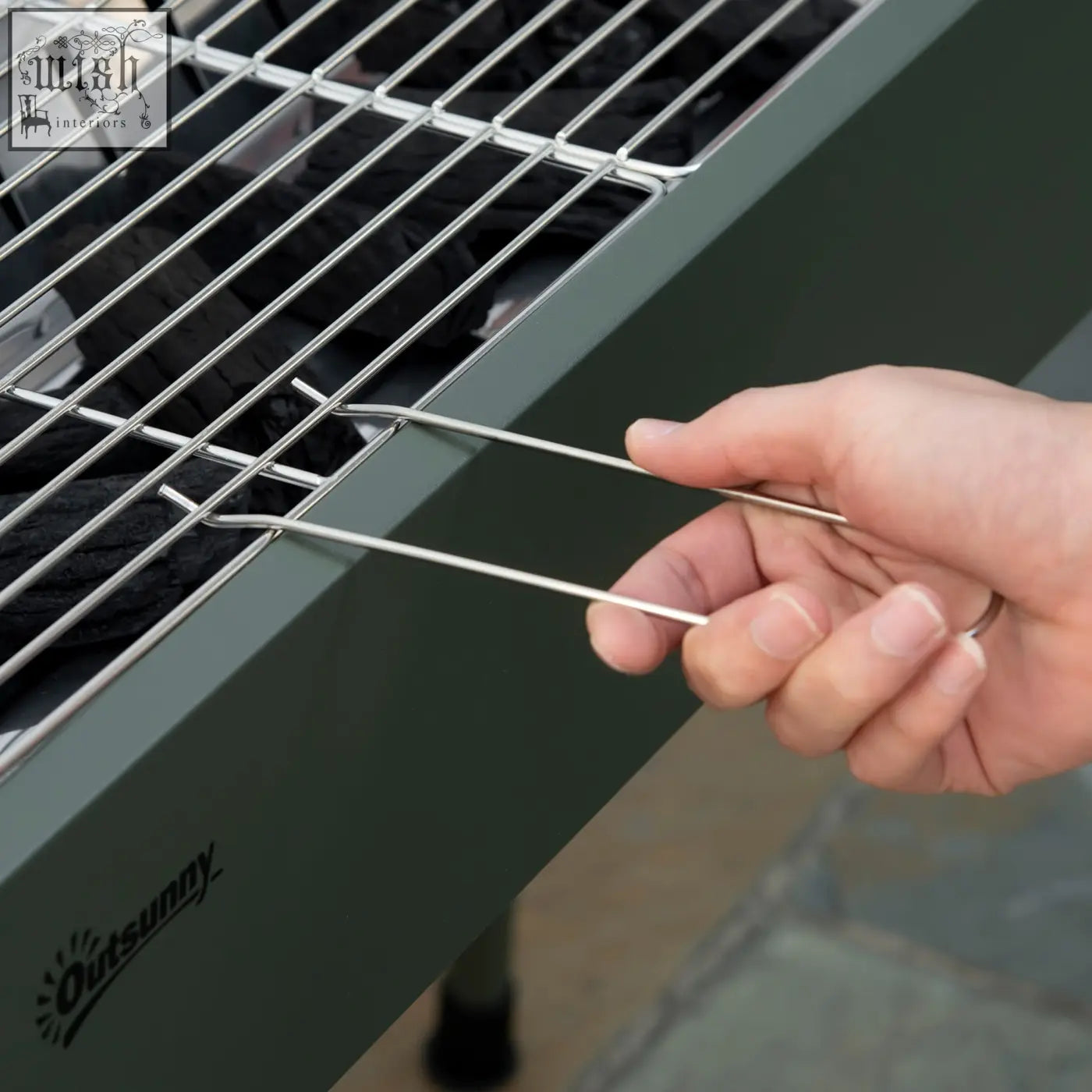 A hand holding a silver metal skewer near the charcoal grate of an outsunny portable stainless steel grill with airflow vents