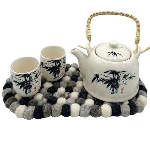 White ceramic teapot with black bamboo design and woven handle on a multi grey oval felt ball centre mat with cups