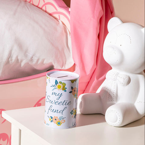 A white cylindrical floral small money tin with colorful patterns and my sweetie fund text next to a teddy bear