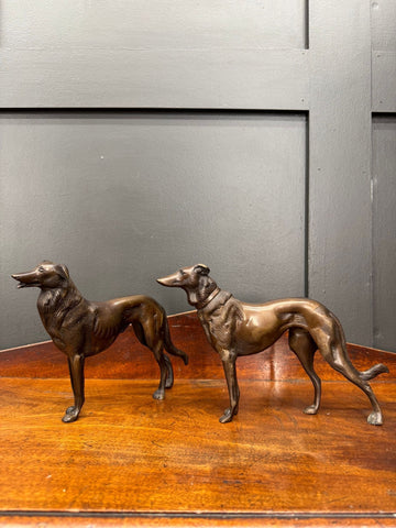 Two bronze greyhound figurines with detailed fur and elegant bodies standing side by side on a wooden surface