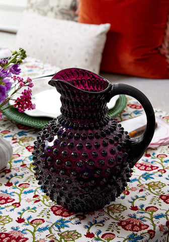 A deep purple hobnail glass pitcher with black handle and rim shown alongside a palm beach cushion cover