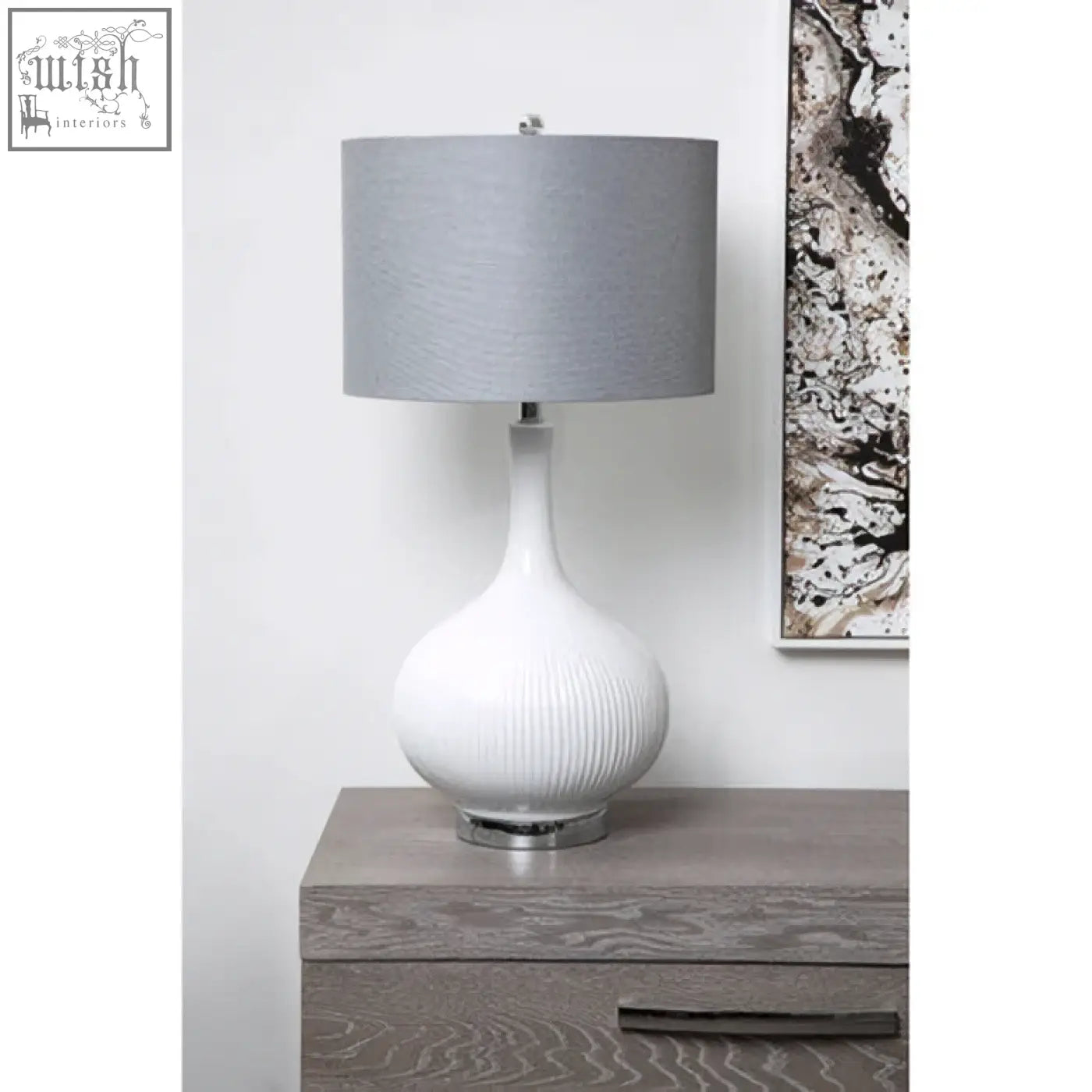 Pandora table lamp is a white ceramic table lamp with a ribbed base, silver metal foot, and gray fabric shade