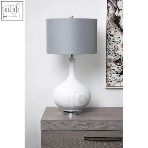 Pandora table lamp is a white ceramic table lamp with a ribbed base, silver metal foot, and gray fabric shade