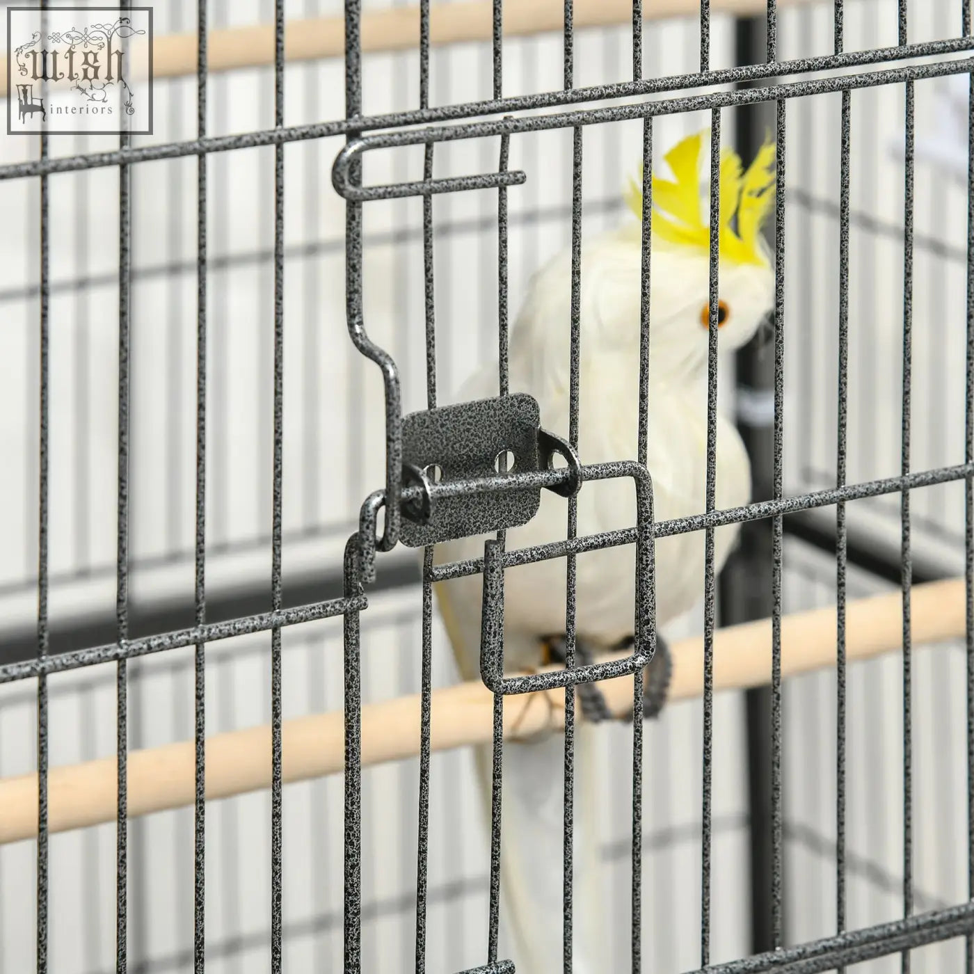 A white cockatiel with a yellow crest inside a dark grey metal bird cage with wooden perches and food containers