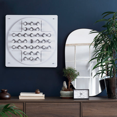 White square wall-mounted peg solitaire art piece with circular grid of recessed round indentations for solo test fun