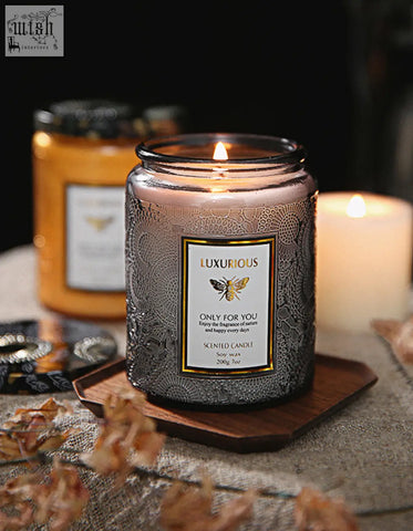 A lit silver glass scented candle with embossed patterns and gold label sits on a wooden coaster perfect as a candles gift