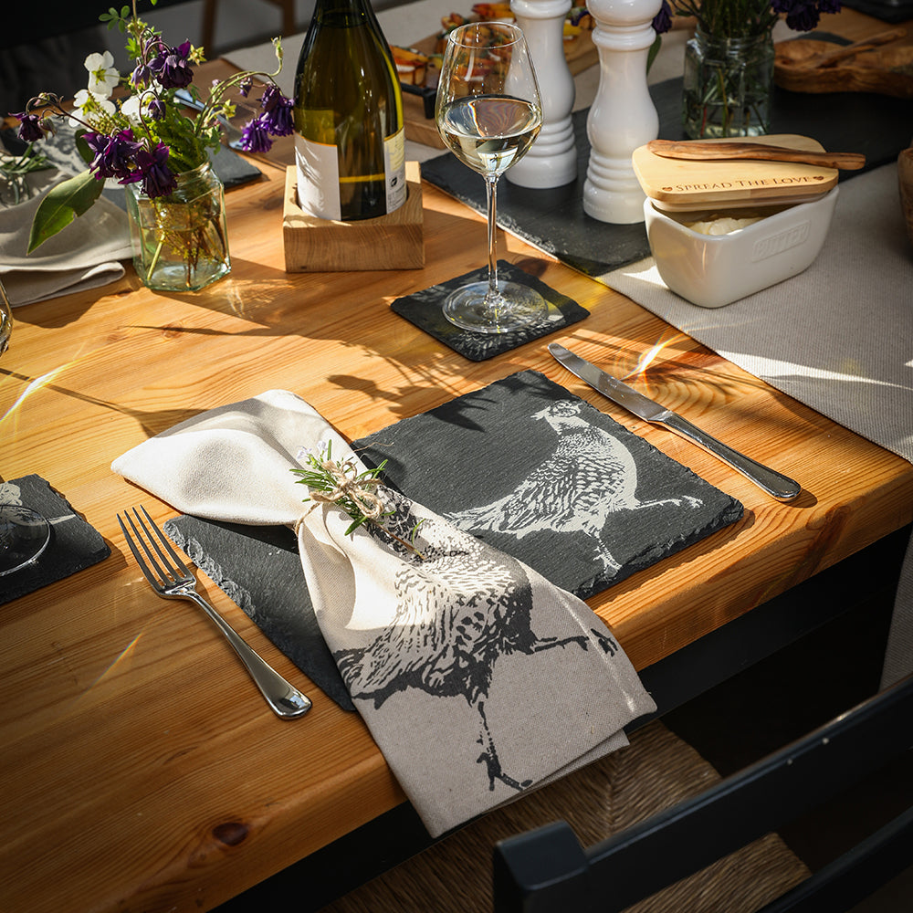 Beige linen napkin with black pheasant design folded on slate place mats set of 2 with fresh herb accent