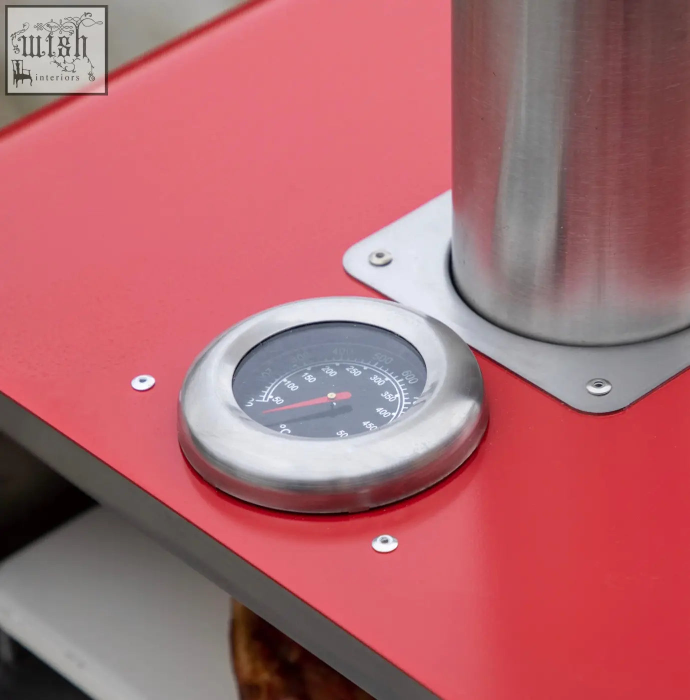Circular stainless steel thermometer with black dial and red needle on a red pizza oven with tall chimney