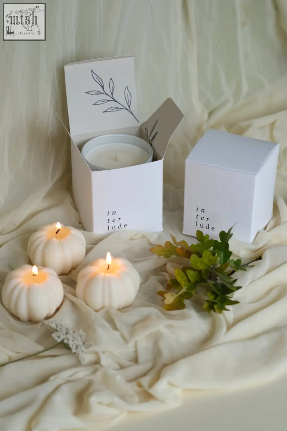 White pumpkin shaped candles with lit wicks in minimalist white boxes featuring delicate leaf art perfect for pumpkin spice decor