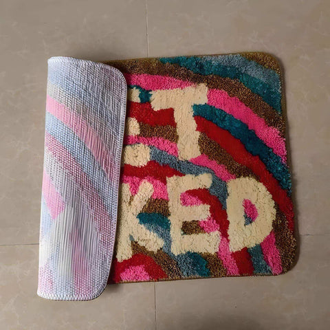 A vibrant naked bathmat with bold rainbow stripes and cream it’s oked lettering in plush shaggy pink teal brown red fibers