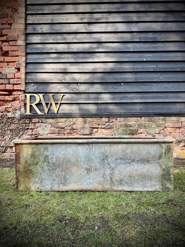 A weathered reclaimed galvanised metal trough planter with rust and moss stains sits on grass by a dark wooden wall