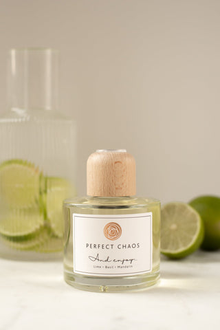 Clear glass reed diffuser bottle with light wood cap and white perfect chaos label showing lime basil and mandarin scents
