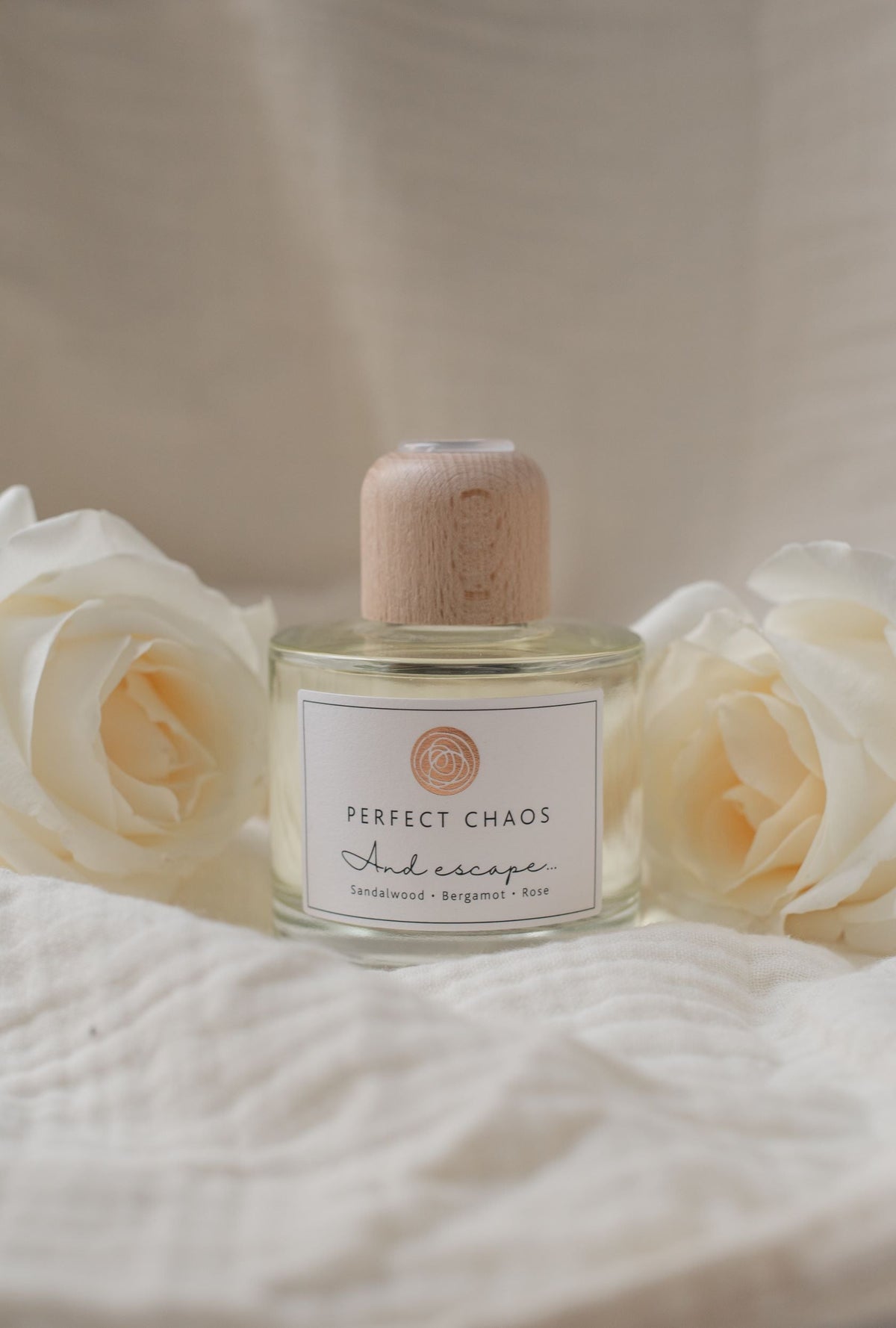 A clear glass reed diffuser bottle with a light wood cap and white perfect chaos label for escape scent