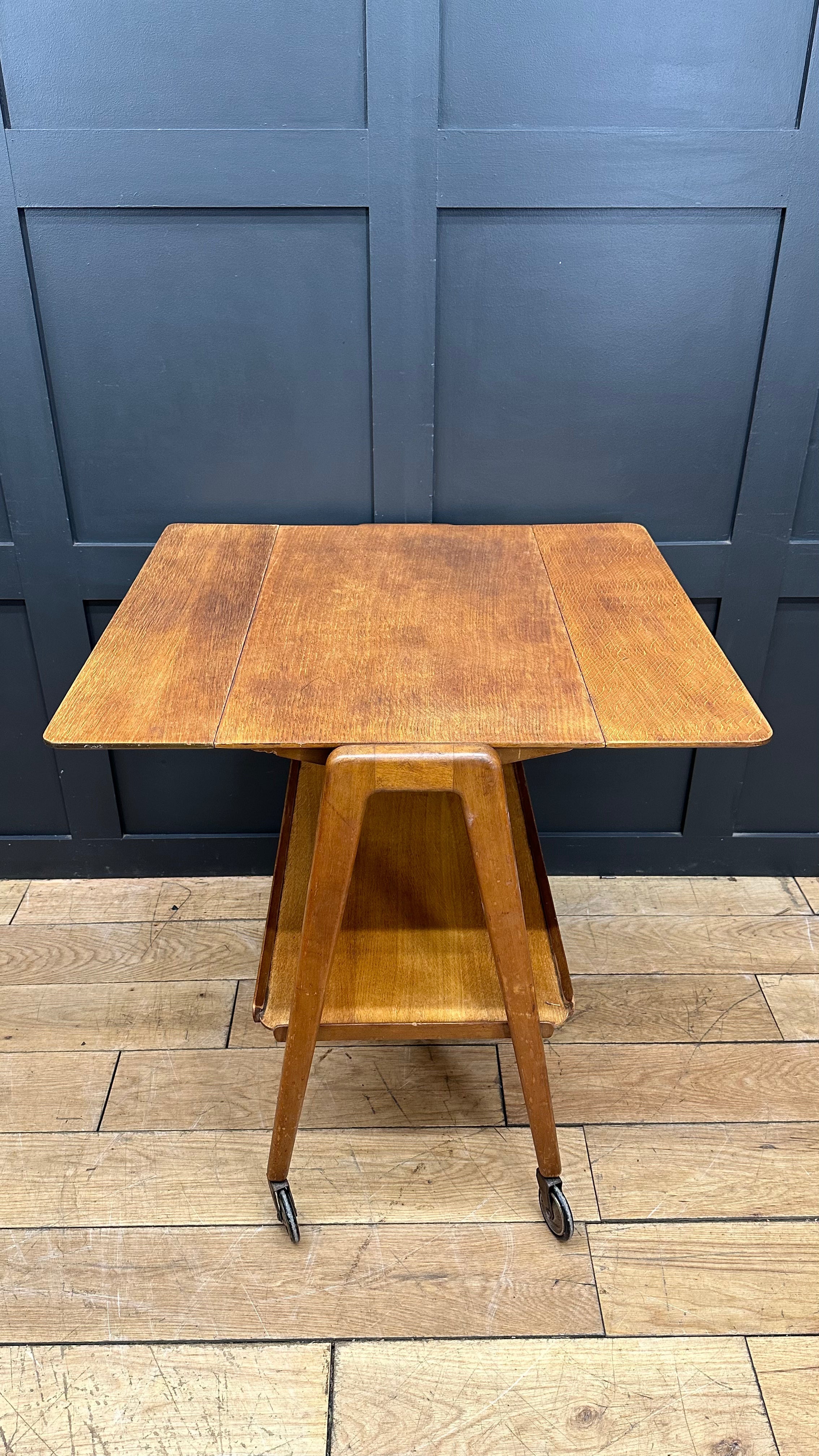 A mid century wooden drop leaf serving cart with warm finish tapered legs two lower shelves and black casters for mobility