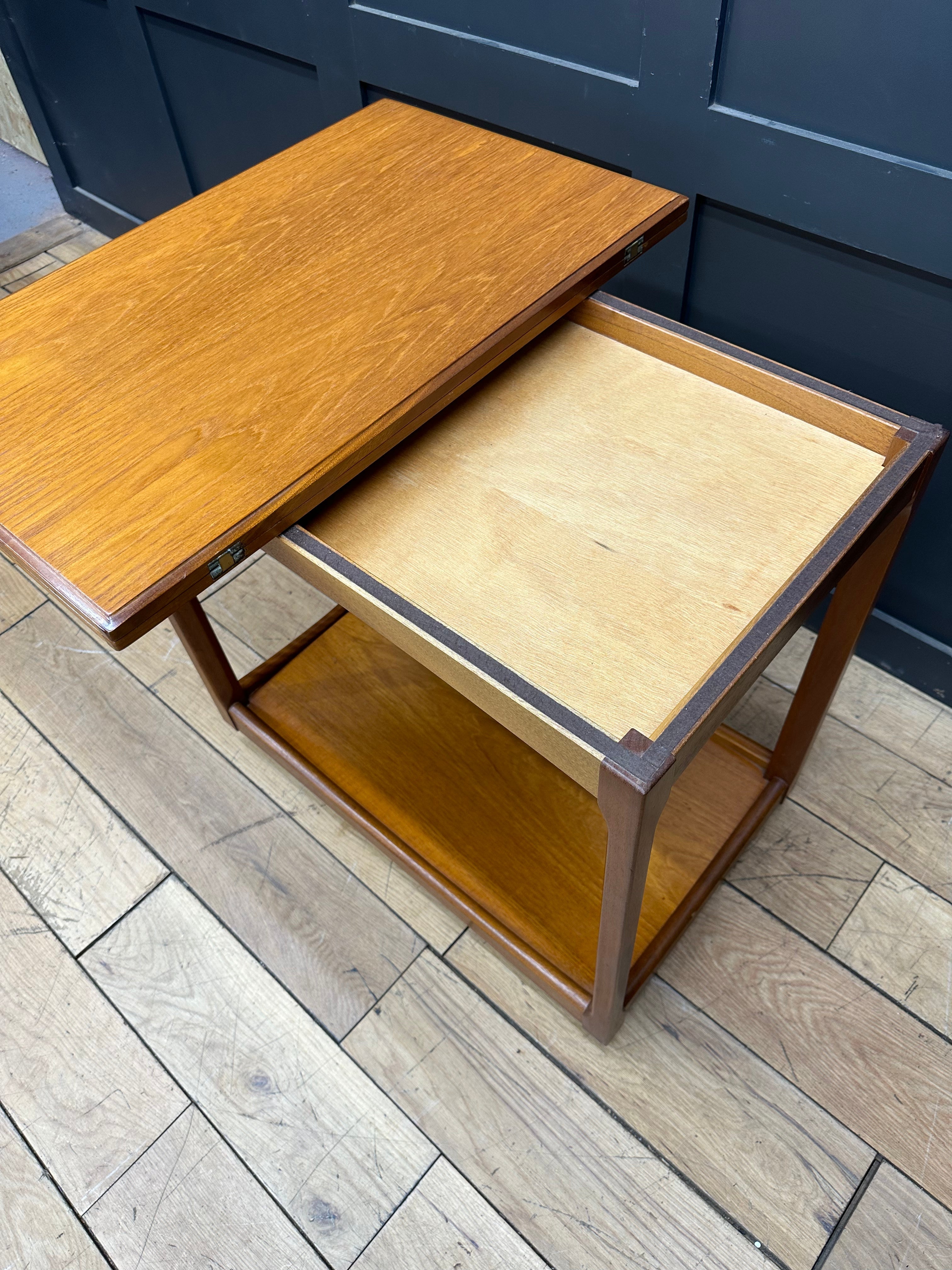 A mid century teak wood coffee table with fold down top and lower shelf in warm golden brown finish for serving