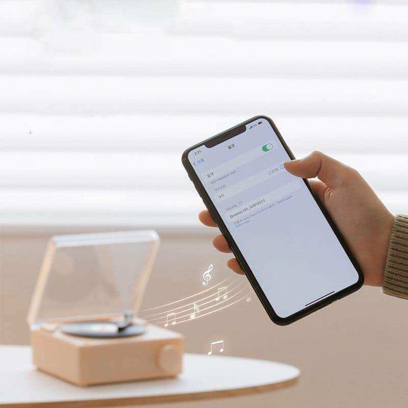 A sleek black smartphone showing a music app in chinese held near a beige retro vinyl wireless bluetooth speaker
