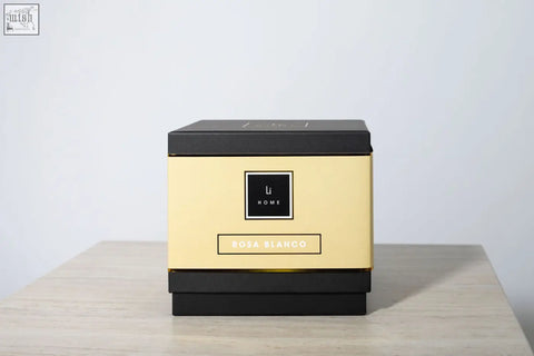 A sleek gold and black square box featuring lu home rosa blanco large candle from a luxury candle collection