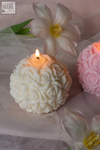 A lit white rose sphere candle with intricate floral detailing and a smooth wax surface glows softly