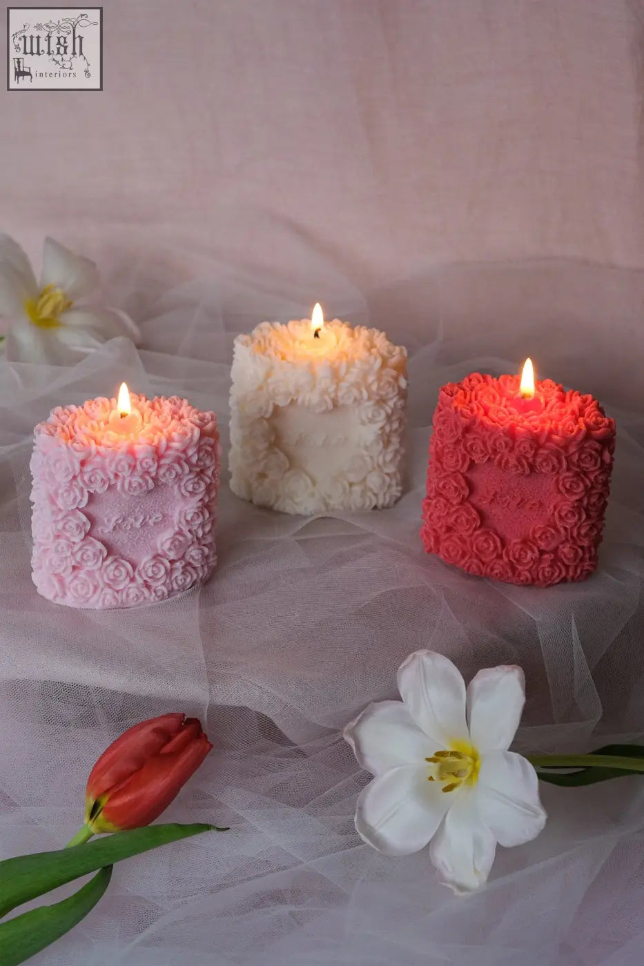 Three lit rose pillar candles in pink white and red with detailed rose patterns on their cylindrical surface