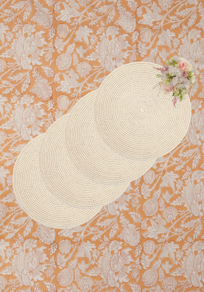 Four cream-colored woven rattan placemats in sand color arranged diagonally on an orange floral surface