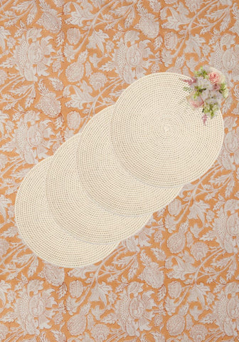 Four cream-colored woven rattan placemats in sand color arranged diagonally on an orange floral surface