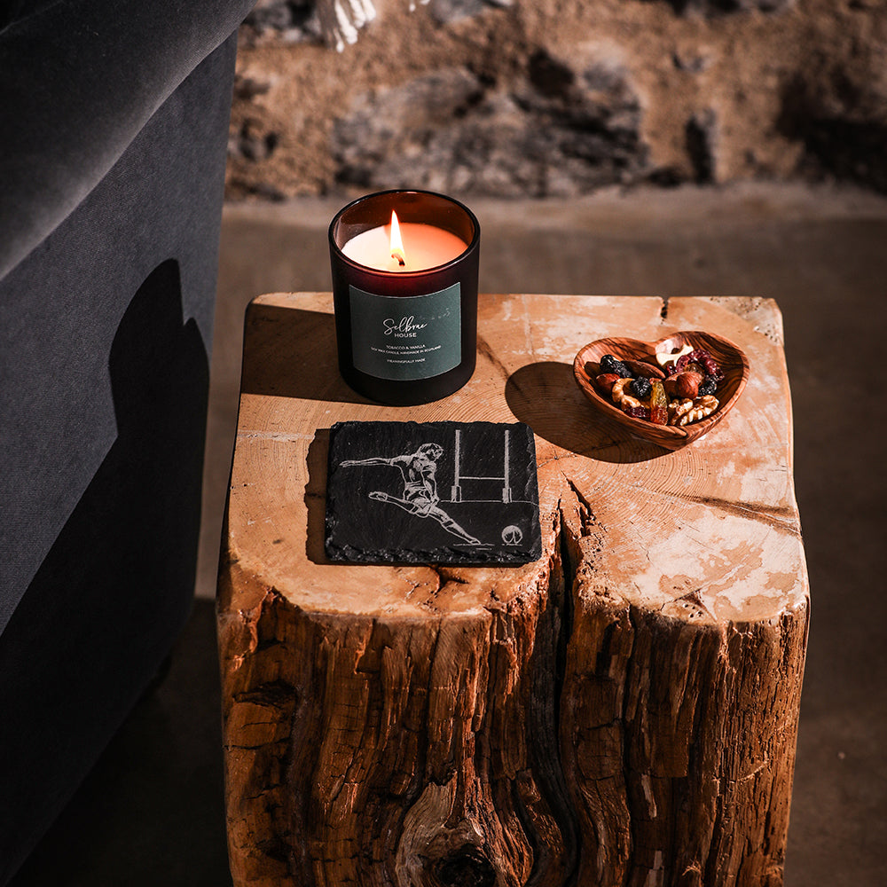 Burning amber glass candle with dark green saffron house label and rugby player illustration on single slate coaster
