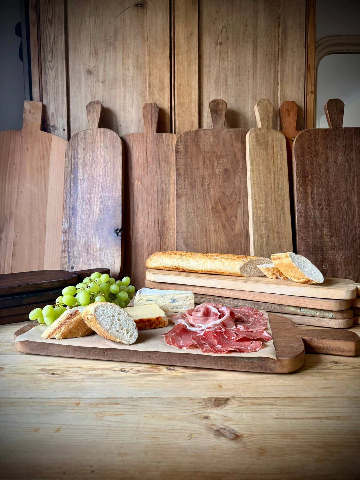Rustic wooden chopping board with natural wood grain and handle displaying cheeses cured meats bread and grapes