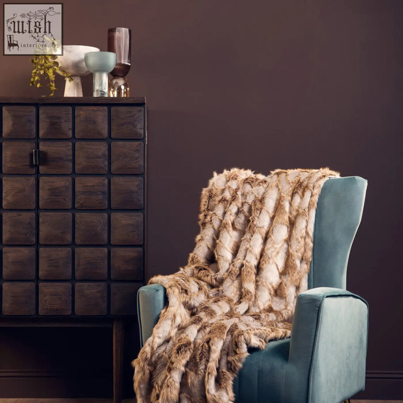 A plush faux fur throw in tan and beige draped over a teal armchair next to a salmo small white marble pedestal bowl