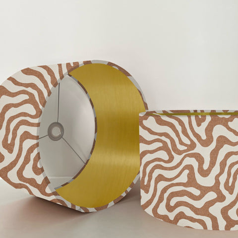 A sandstone wiggle drum lampshade with bold brown and white waves and a metallic brushed gold interior lining