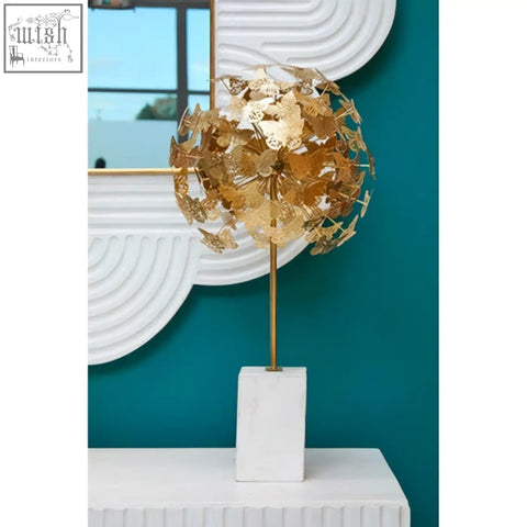 Golden metallic dandelion sculpture with intricate petals on a white marble base next to a mango wood wall mirror