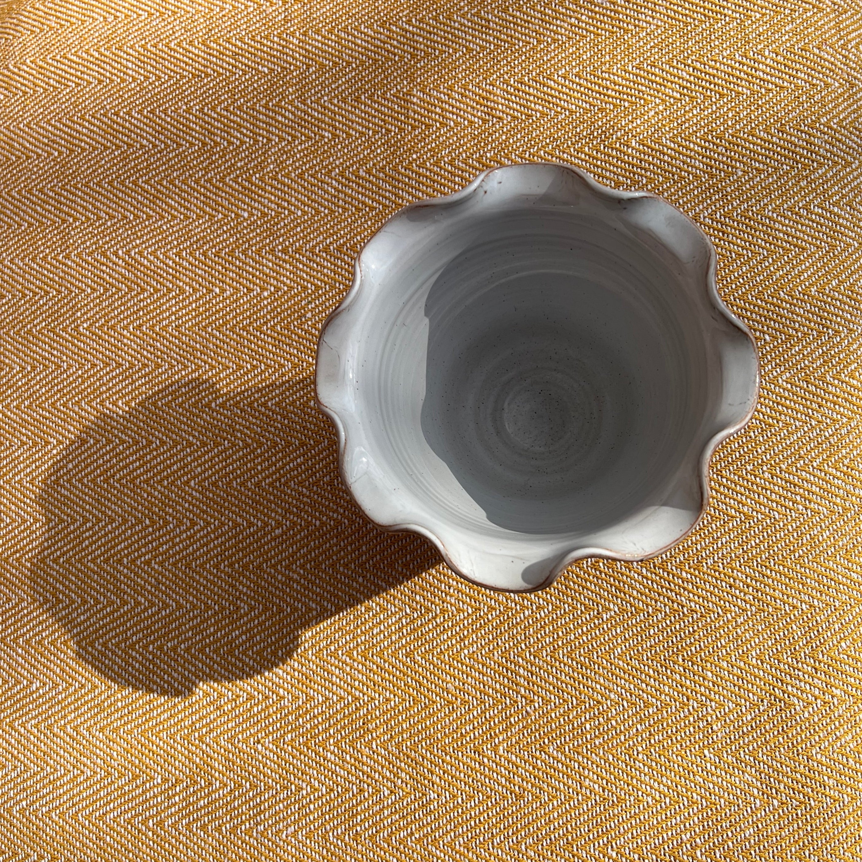A small light gray ceramic scalloped bowl sitting on textured mustard yellow fabric in scalloped brown vase pot style