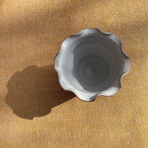A small light gray ceramic scalloped bowl sitting on textured mustard yellow fabric in scalloped brown vase pot style