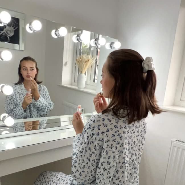 A woman in a white and black floral pajama set applies lipstick at a vanity with bright lights on the scarlett hollywood mirror 100x70cm