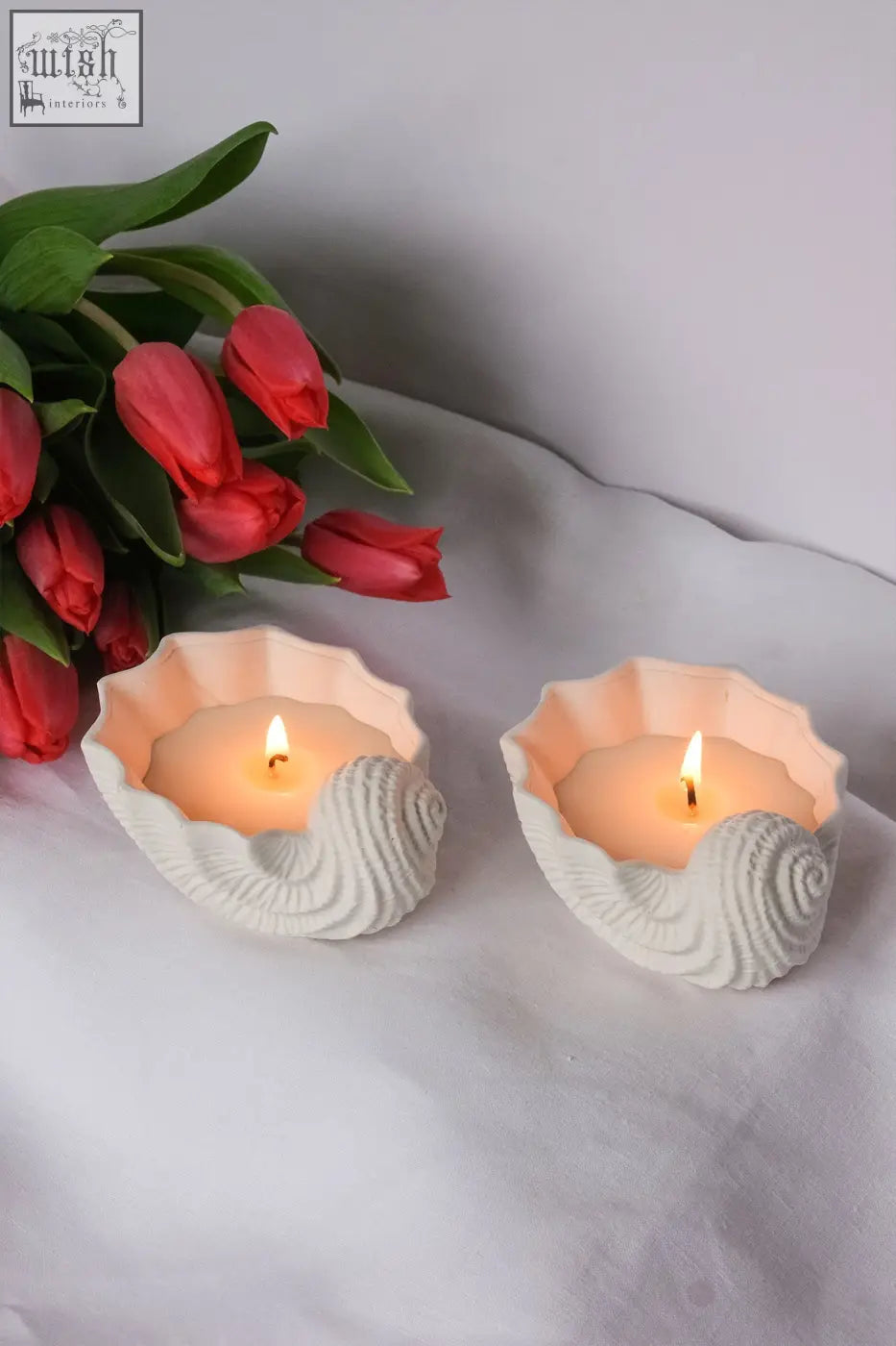 Two white seashell shaped candle holders with lit ylang ylang scented candles inside