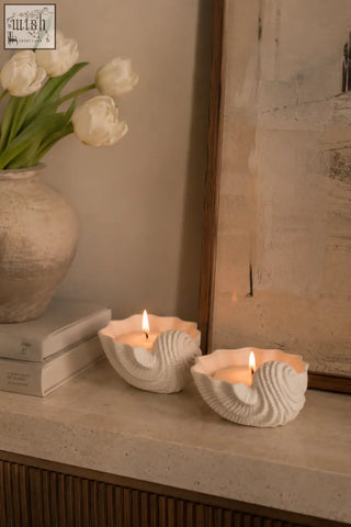 Two white ceramic seashell shaped candle holders with lit ylang ylang scented candles inside