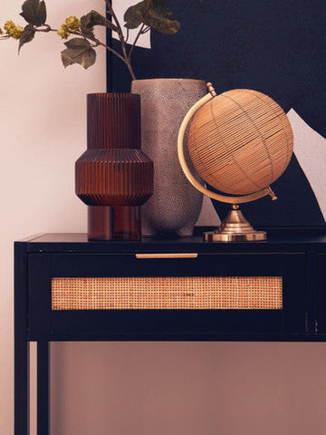 A woven rattan globe with brass stand and metallic rim displayed on the sherman black wood rattan two drawer console table
