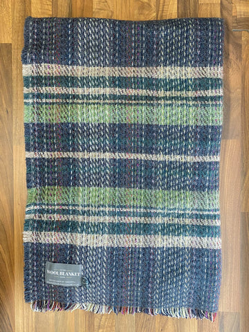 A cozy small checked random recycled wool blanket with a navy, green, and beige plaid pattern and fringed edges