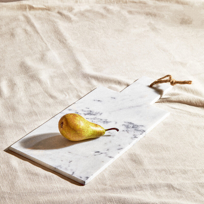 A small white marble chopping and serving board with gray veining and rope handle holding a golden pear