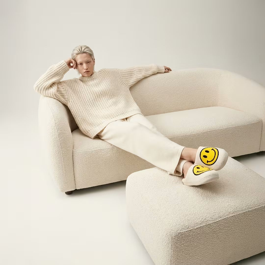 Cream-colored smiling slippers with bright yellow smiley face and plush faux fur texture slip on design