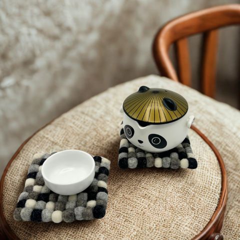 A charming white ceramic panda teacup with a golden lid sitting on a black and gray square felt ball coaster set