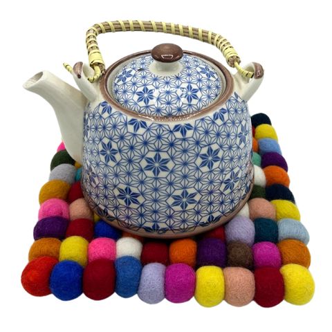 White ceramic teapot with blue floral patterns and bamboo handle on a colorful multi colour square felt ball placemat