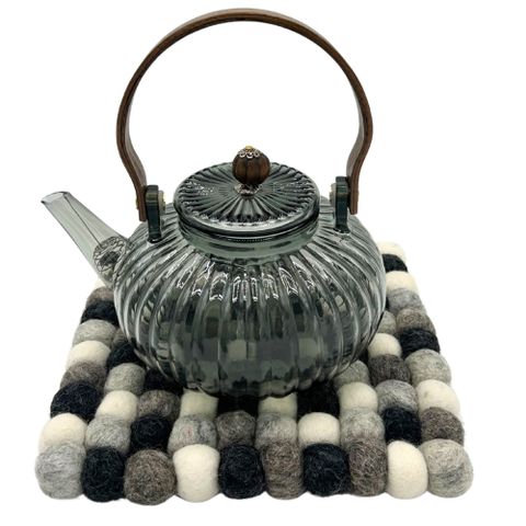 A dark gray ribbed glass teapot with wooden handle sits on a square felt ball placemat made of multi grey wool balls