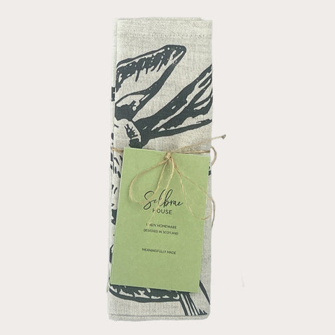 A neatly rolled light gray linen napkin with a bold black bird graphic tied with twine from selfridges house