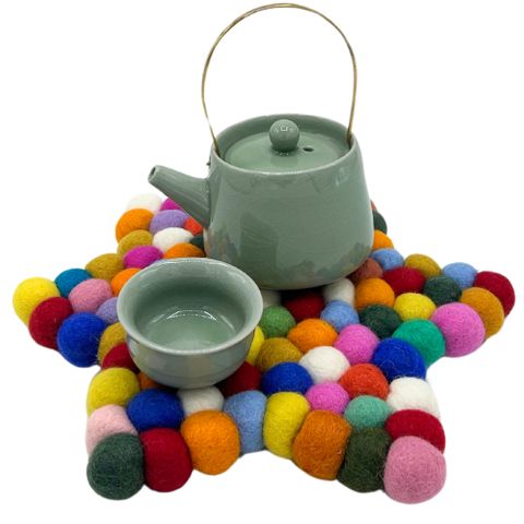 A light green ceramic teapot sits on a multicolor star shape felt ball centre mat made of fuzzy pom-poms