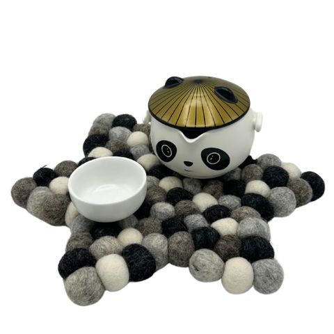 A charming white ceramic teapot with gold leaf lid and black eyes sits on a star shape felt ball centre mat in multi grey