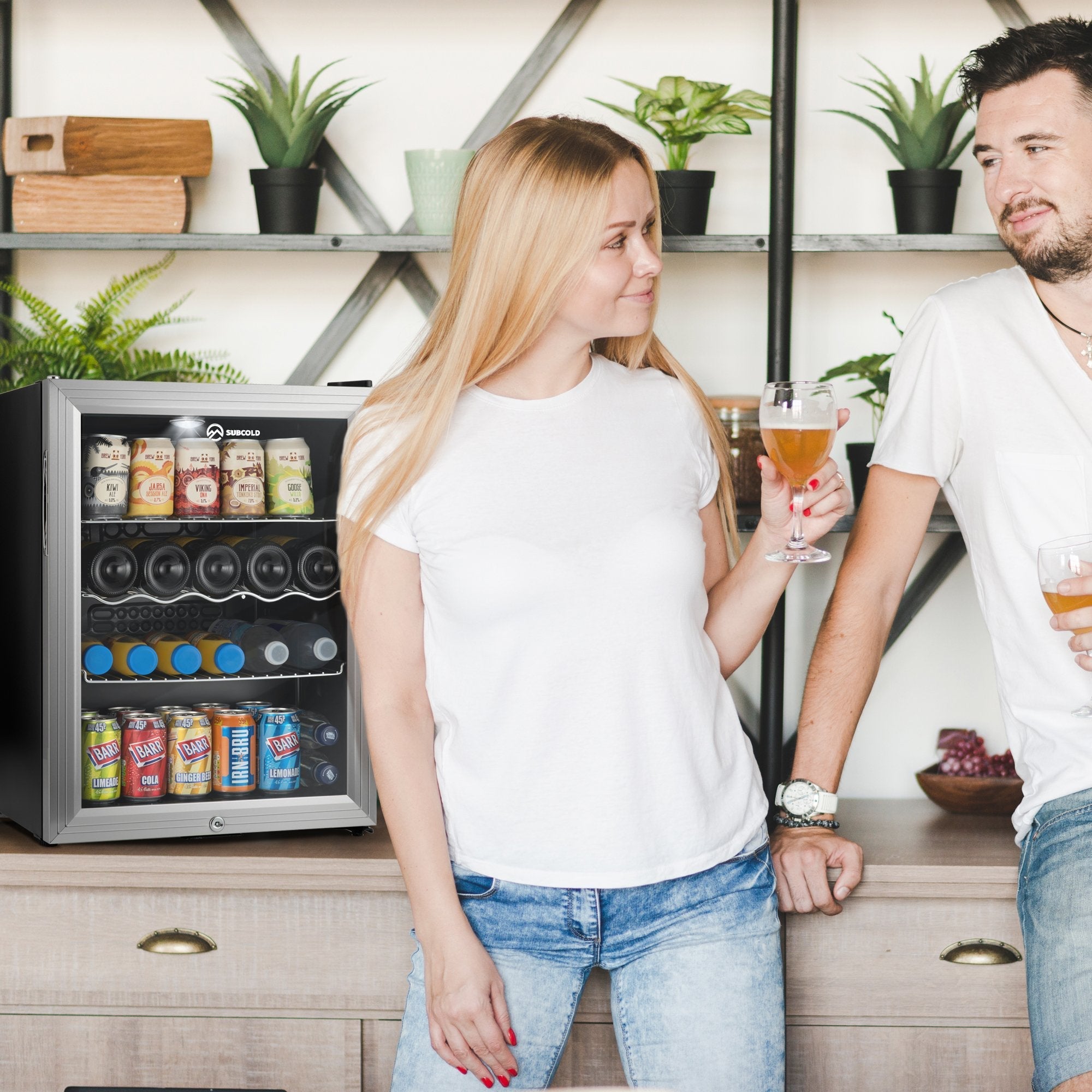 Silver-framed subcold super65 drinks fridge with glass door and black shelves holding assorted canned drinks