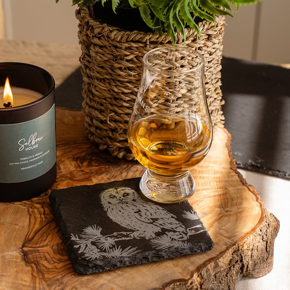 A clear glass whisky tasting glass with amber liquid on a dark slate coaster featuring an engraved tawny owl design