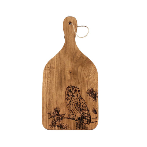 A light brown oak paddle cutting board with a laser engraved tawny owl on pine branches and a hanging loop